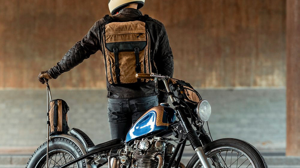 Motorcycle rider with brown backpack standing behind a Yamaha chopper styled motorcycle.
