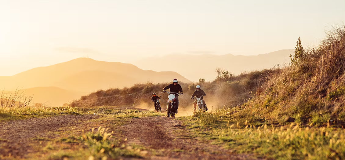 Three motorcyclists ride along a dusty gravel road through rolling hills at golden hour. Dust trails rise behind the motorcycles as mountains stretch across the horizon, capturing a sense of freedom, adventure, and camaraderie.