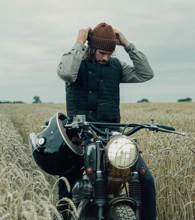 Man dressed in shirt and west sitting on a Kawasaki W650 motorcycle in the middle of wheat field.