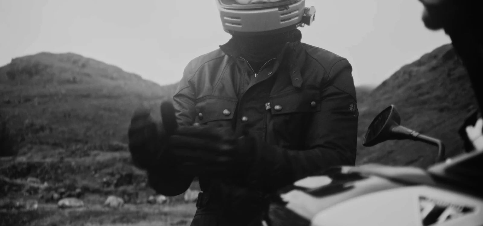 Motorcyclist wearing a classic Belstaff motorcycle jacket and helmet in a rugged mountain landscape.