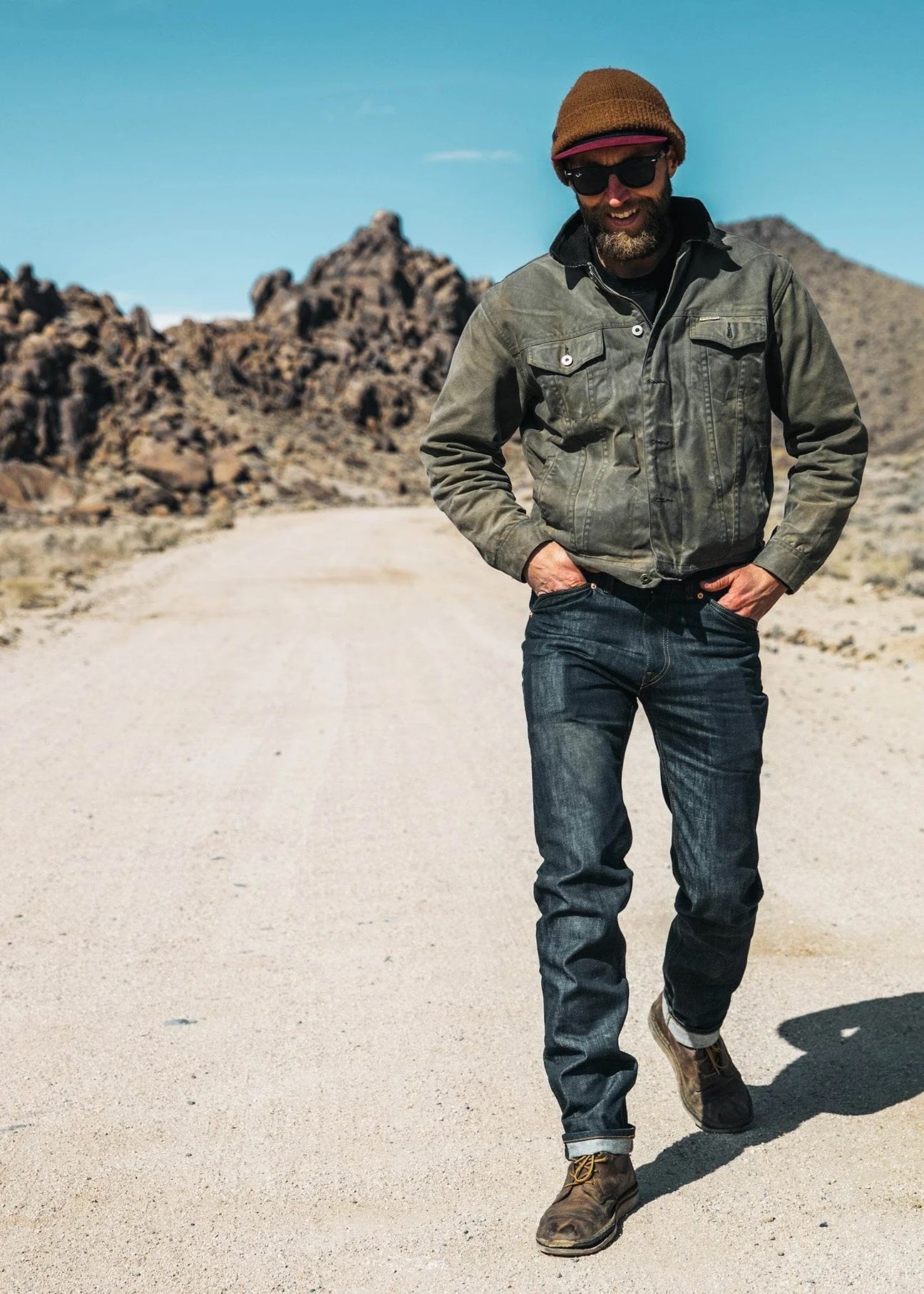 Man wearing sunglasses and wearing denim pants and jacket walking on a gravel road with hills in the background.