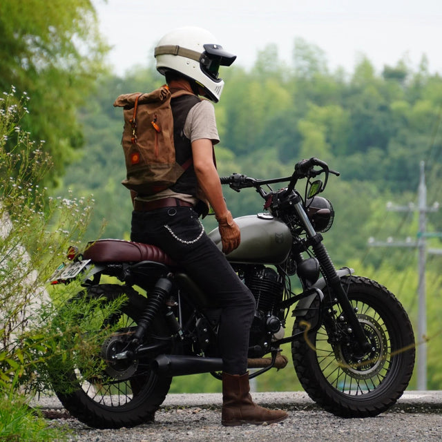 Man wearing t-shirt, jeans, white helmet, motorcycle goggles and waxed cotton backpack sitting on a scrambler style motorcycle.