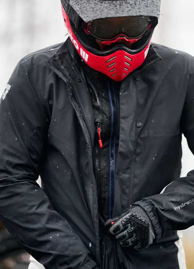 Motorcycle rider in red motocross helmet and goggles is putting on a rain jacket.