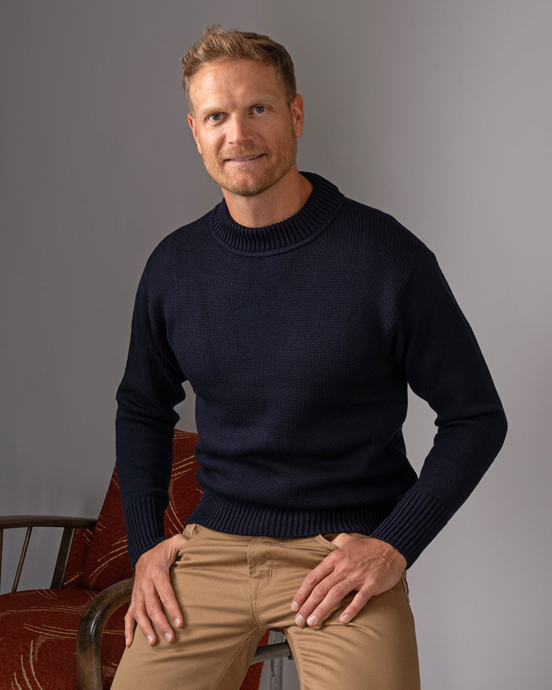 Man dressed in light brown pants and knitted navy sweater while leaning against a chair.