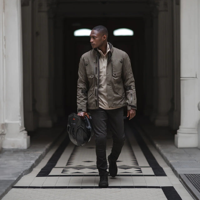 Man in motorcycle jacket holding black motocross helmet walking on black and white tiled floor.