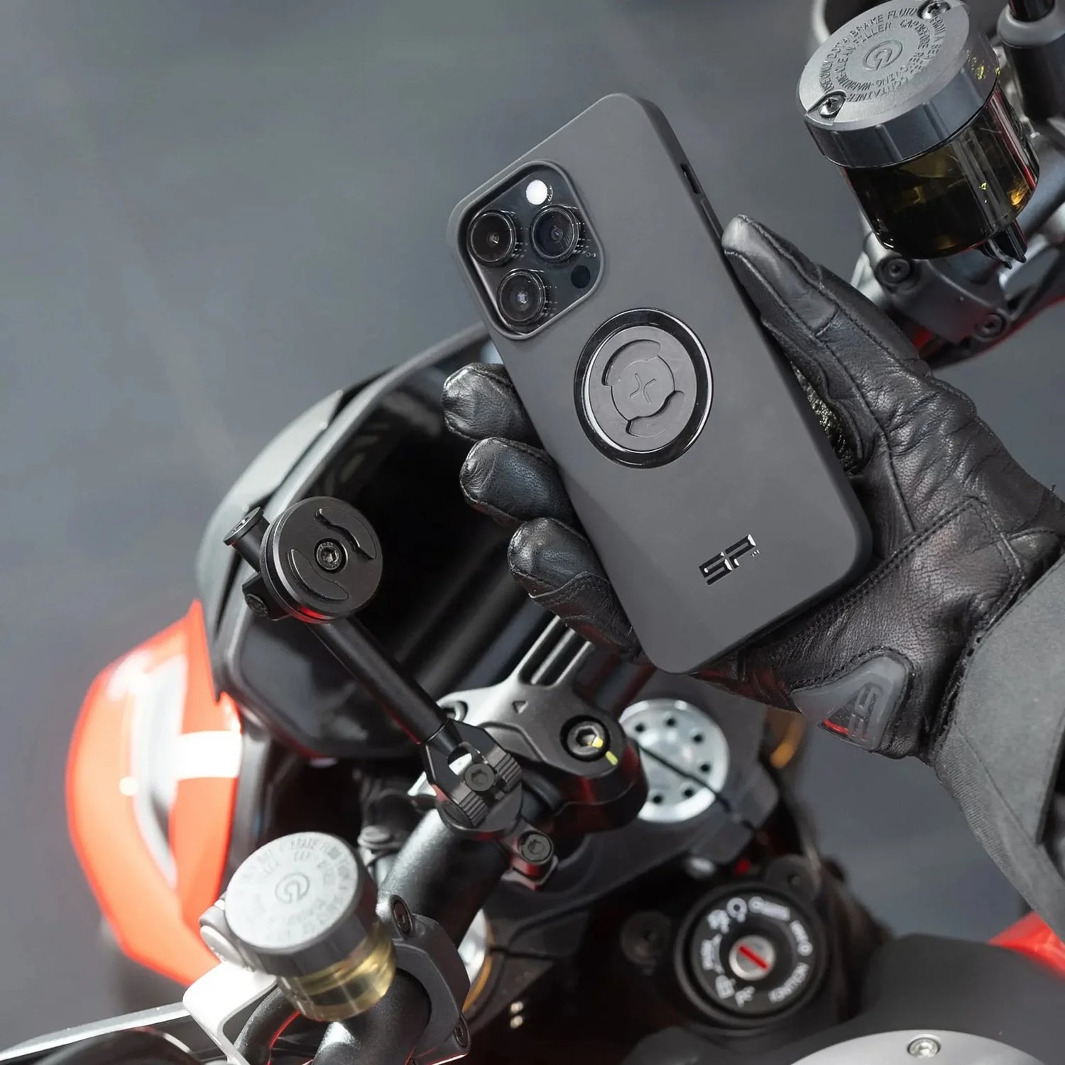 Leather gloved hand holding a mobile phone with a case for mounting it on a motorcycle handle bar.