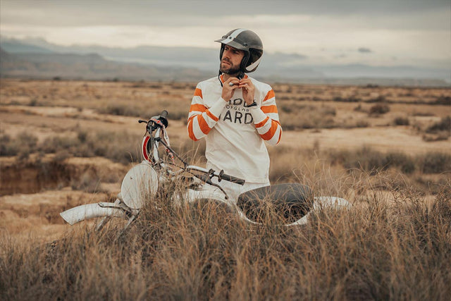 Man in open faced helmet and white sweater with red striped arms stands behind a vintage scrambler motorcycle. Backdrop is an open countryside with mountains in the far distance.