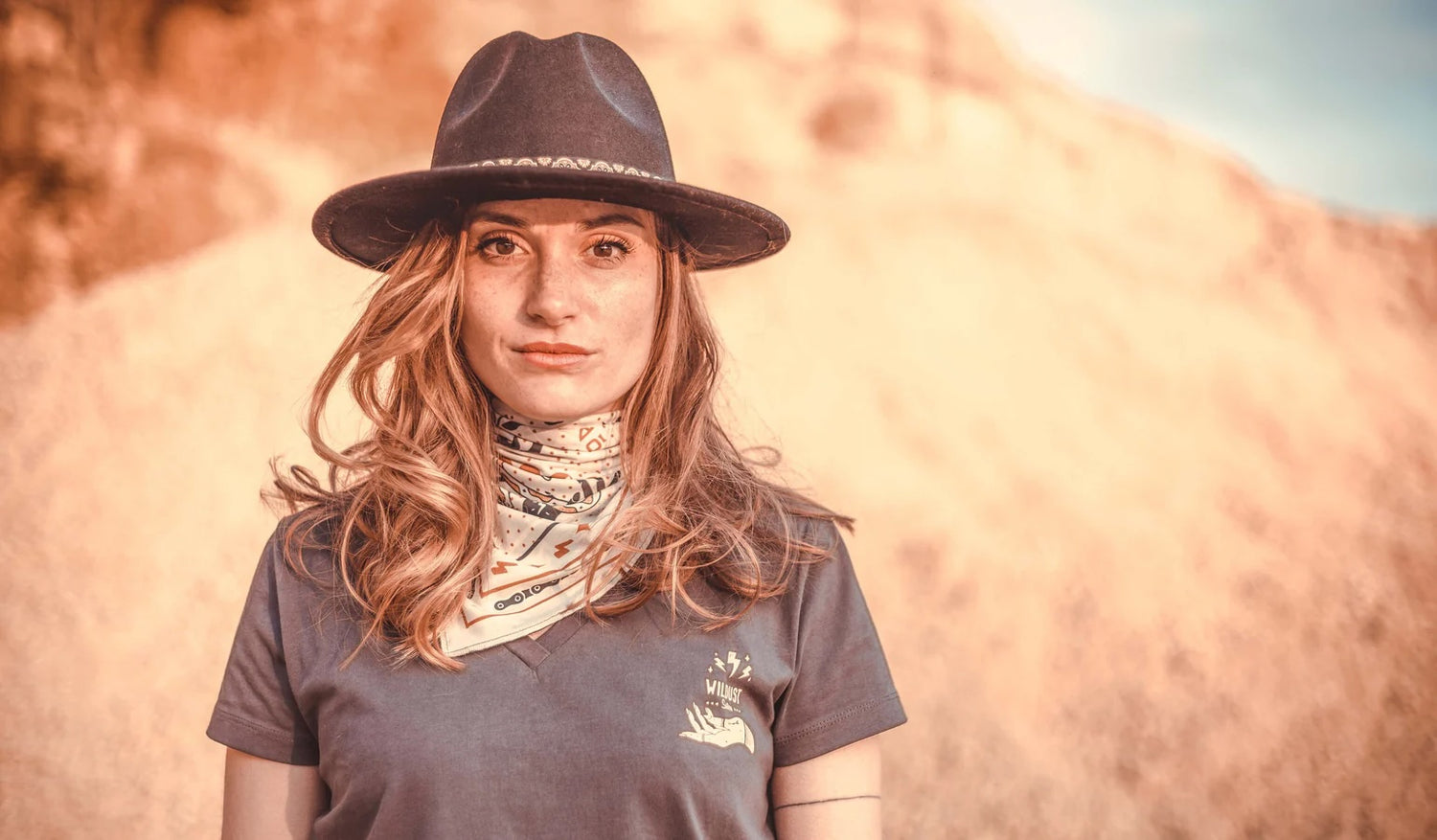 Woman with long hair dressed in a fedora hat, scarf and v-neck t-shirt.